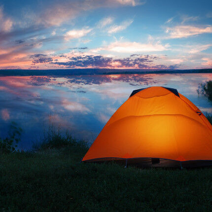 Internally lit orange tent on shore of lake under dramatic sunse
